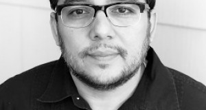 Black and white headshot of Matthew Paul Olmos, a Mexican-American man looking studious with rimmed glasses and a collar shirt with a plain background.