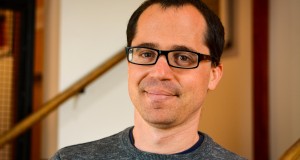 Headshot of Andrew Rosendorf, wearing a black sweater, glasses, and smiling on the stairwell of the Playwrights' Center