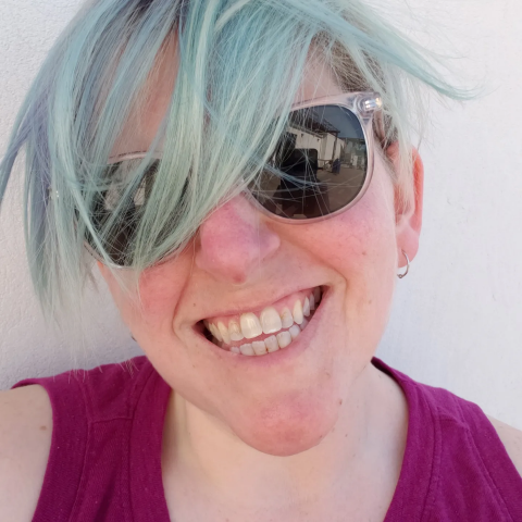 Headshot of C. Meaker, a queer white person with blue hair smiles at the camera. They wear a pink tank top on a sunny day with a white background.