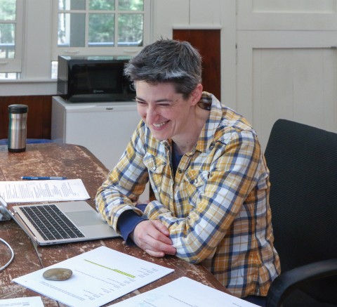 Headshot of L M Feldman, looking down with a bashful smile, sitting at an old wooden desk covered in papers and a laptop; L is white, is wearing a flannel yellow-and-blue-plaid shirt, and has super-short-cropped brown hair that’s graying. :)