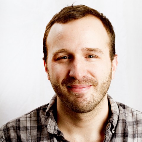Paul Kruse, a white man with short brown hair and stubble wearing a grey flannel shirt, smiles in front of a white background.