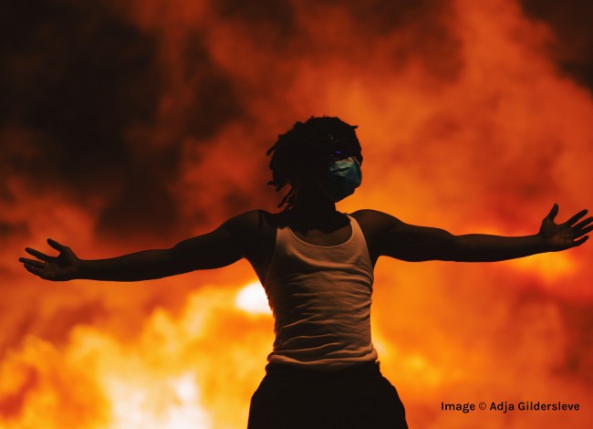 copyright Adja Gildersleve: A black man in a white tank top stands with his arms stretched out wide. He stands in front of  a sky filled with flames.