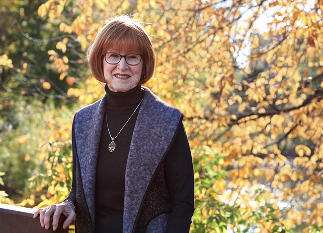 Ginger Wilhelmi smiles at the camera. She is wearing glasses, a black turtleneck, and pendant necklace. The background of fall foliage, with yellow, red, and green leaves compliments her auburn hair. 