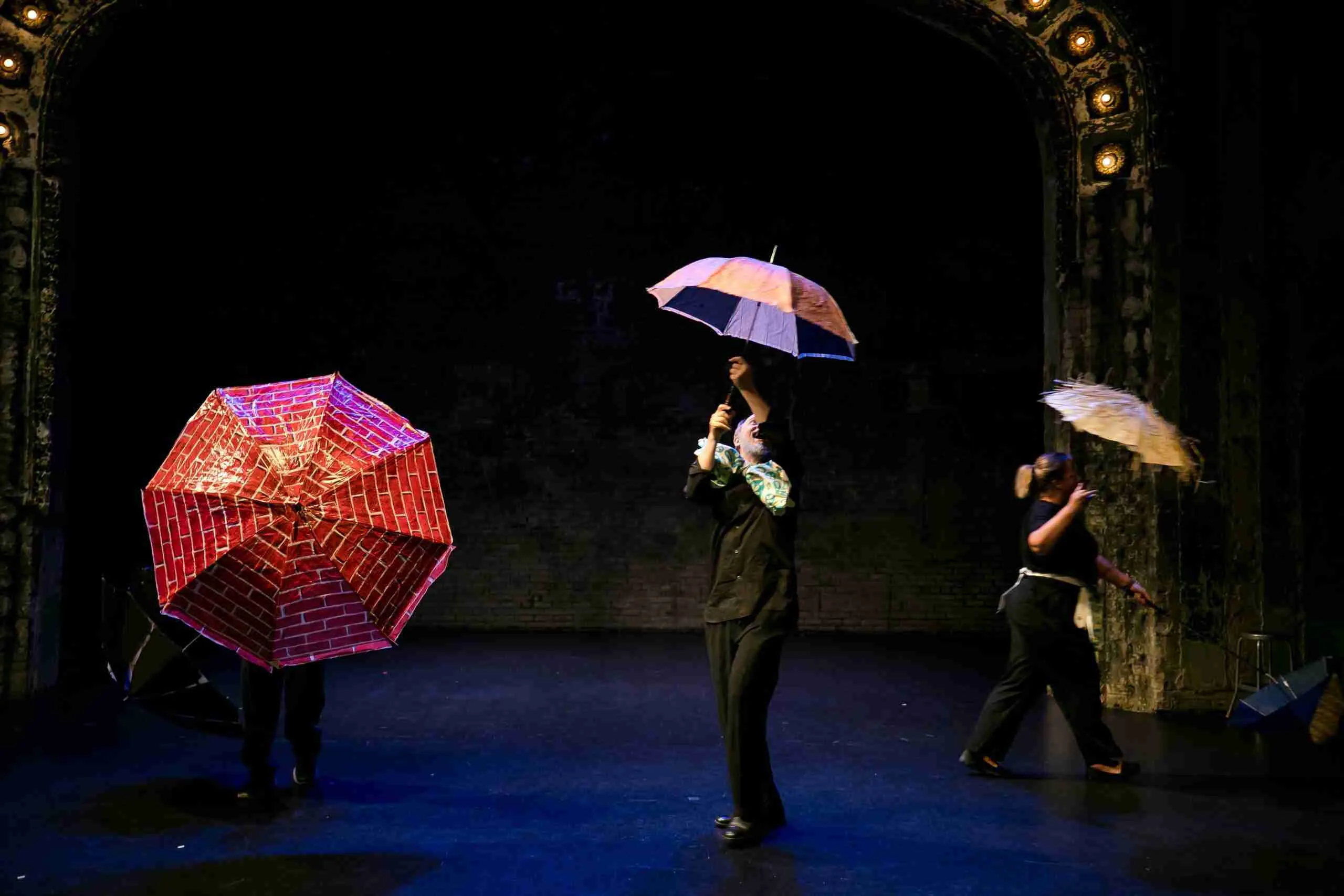 Three actors dance on stage holding umbrellas.