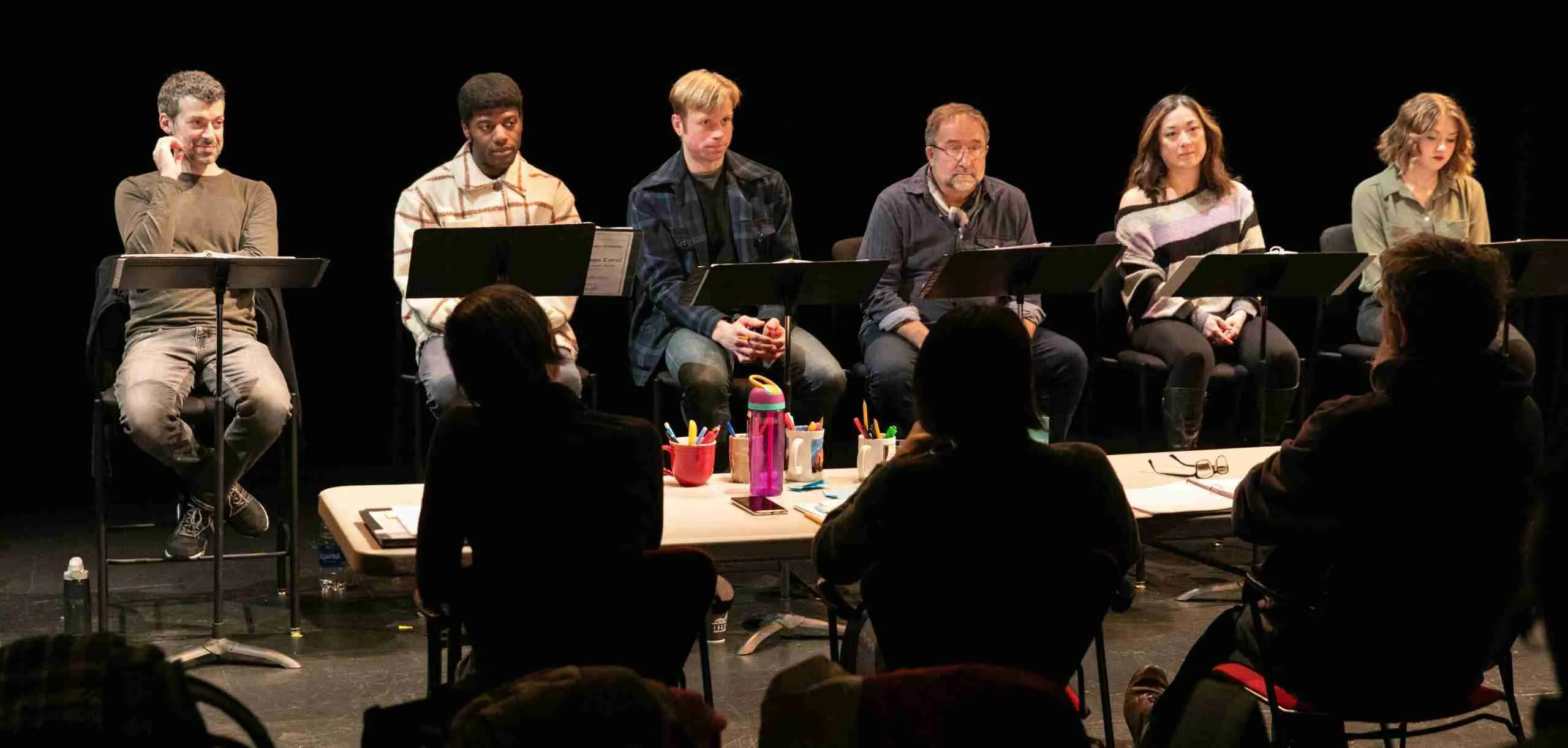 A playwright and her collaborators sit at a table facing several actors at music stands to give notes.